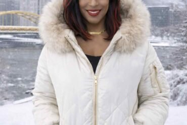 Single in the Steel City 2026 winter portrait of a woman in Pittsburgh wearing a silver bomber jacket with a white fur hood, standing outside during heavy snowfall with a confident smile and red lipstick.